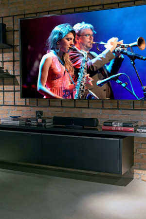 Preso na parede de tijolo, o TV de 85” fica na frente de uma grade metálica, que serve de apoio para as prateleiras.