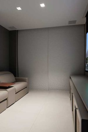 Na pequena sala de home theater, tela de 100”, projetor 4K e revestimento acústico atrás do sofá.