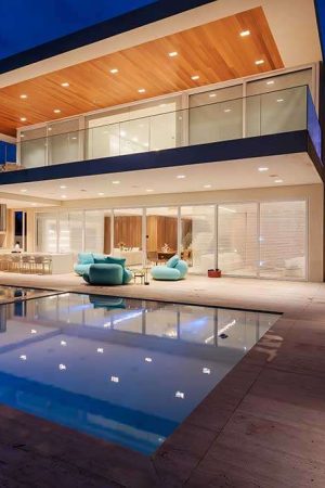 Vista ampla da área externa, com a piscina refletindo o azul do céu e as luzes em volta da casa.